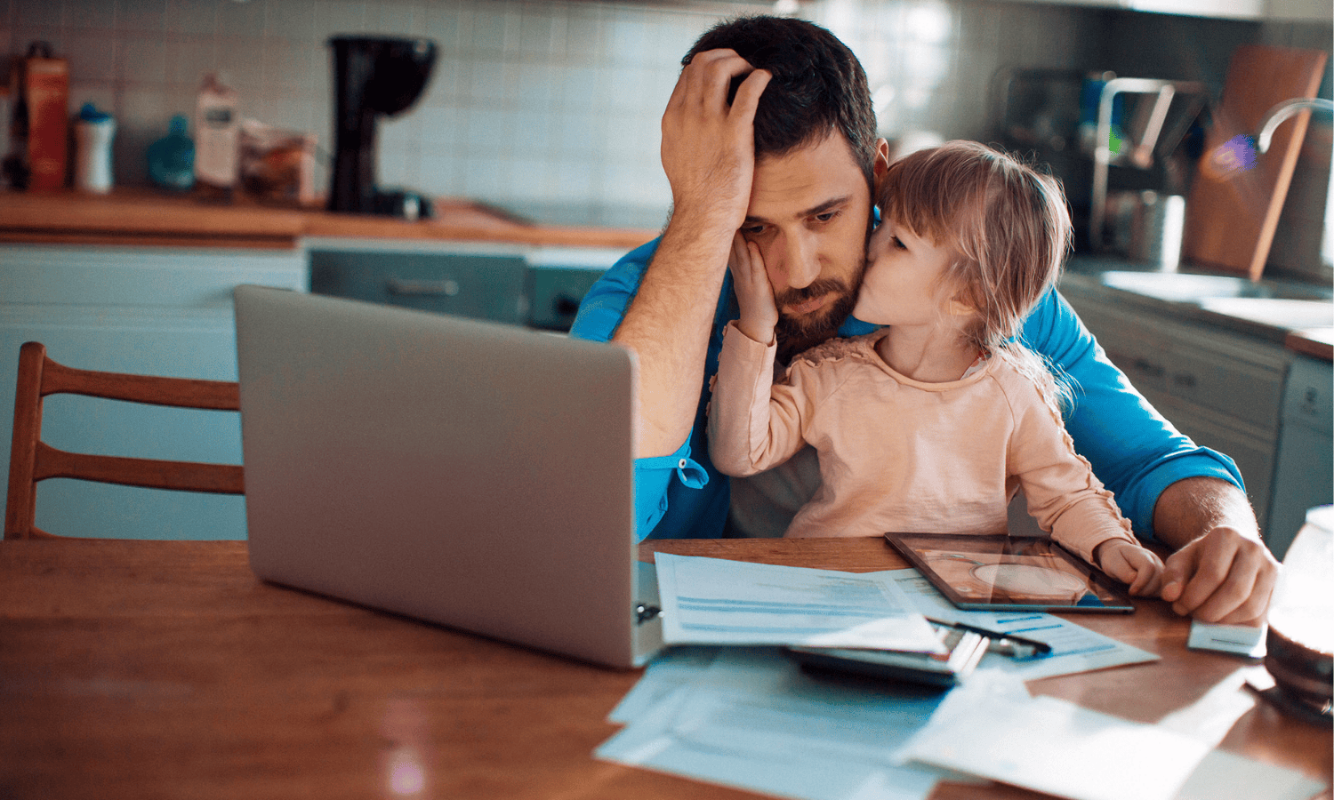 Dad looking distressed in front of his computer and bills with his daughter on his lap.