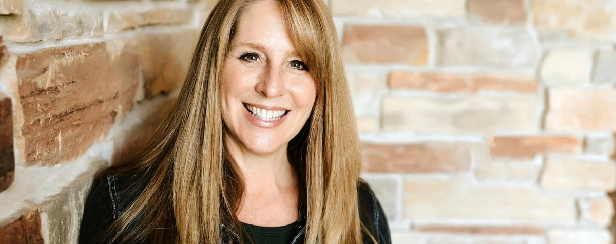 Photo of speaker Holly Schneider wearing a black t-shirt and black jean jacket. Holly is leaning against a cream brick wall.