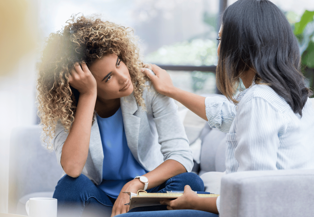 A woman rests her head on her hand while another woman reaches out to touch the first woman's shoulder.